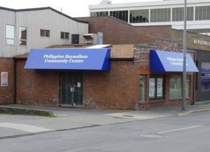 Photo: A corner of a brick one-story building, with overhanging blue signs on two sides that say: Philippine Bayanihan Community Centre.