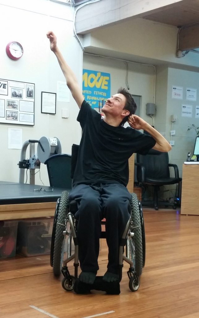 Kristian is turning his head and looking up to the top left corner of the photo, with a smile. He has one arm diagonally up to the left and his other elbow is out to the right with his hand by his ear. He is wearing a black t-shirt and dark pants and is seated in his manual wheelchair. There is some gym equipment in the background.