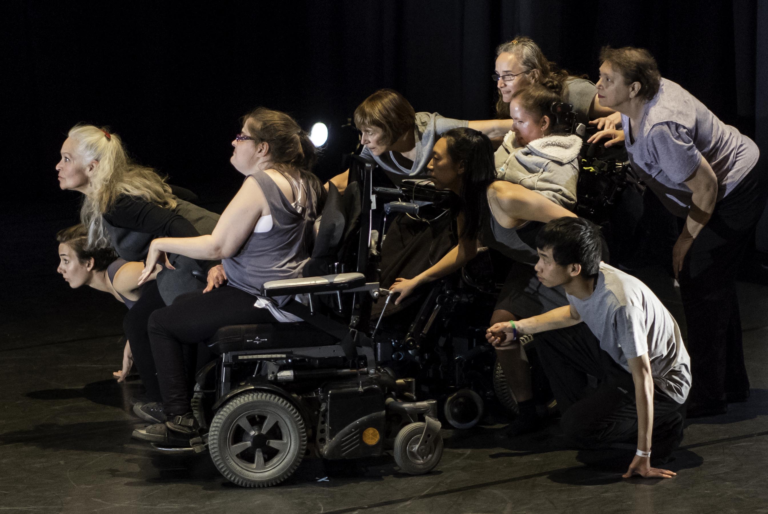 Photo of nine dancers huddled together, crouched and seated, attentively staring to the left.